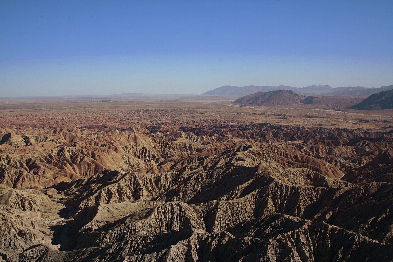 Aerial view of Anza-Borrego badlands showing densely eroded tan and brown ridges stretching toward a flat desert valley and distant mountains under clear blue sky