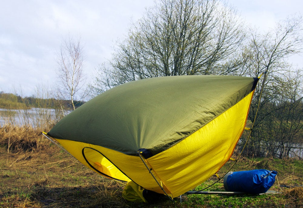 Yellow and olive green dome tent billowing in wind beside a calm river, bare early-spring trees behind, blue dry bag on ground nearby